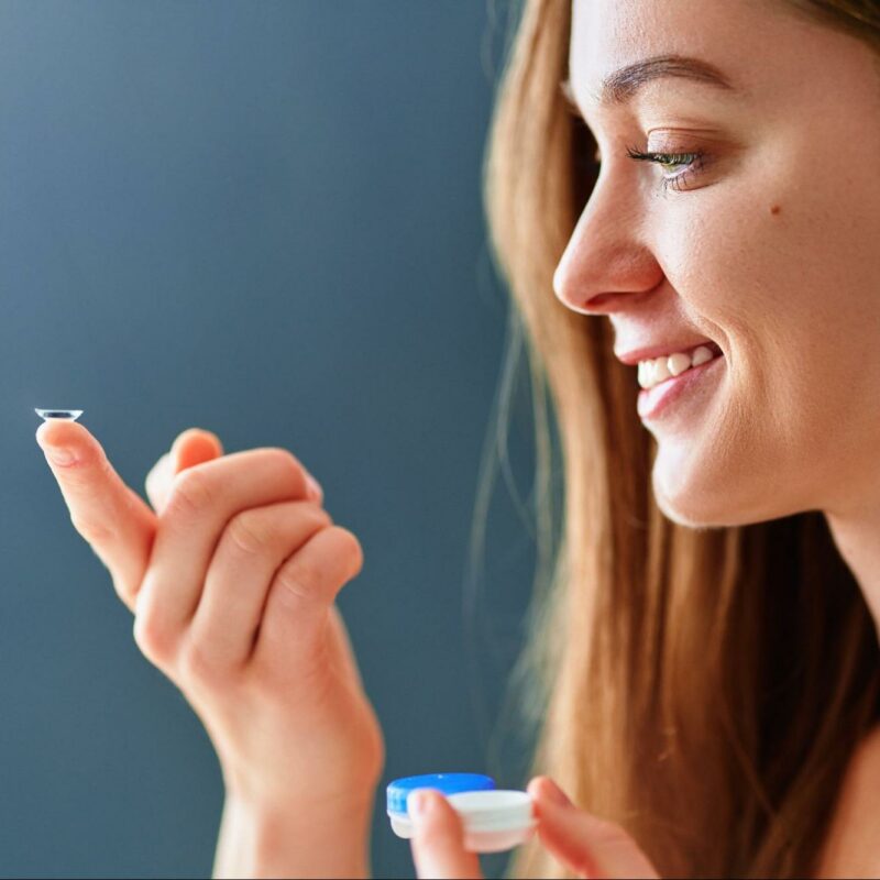 A smiling woman holding a contact lens on her fingertip, ready to put it in her eye, with a contact lens case held in the other hand.
