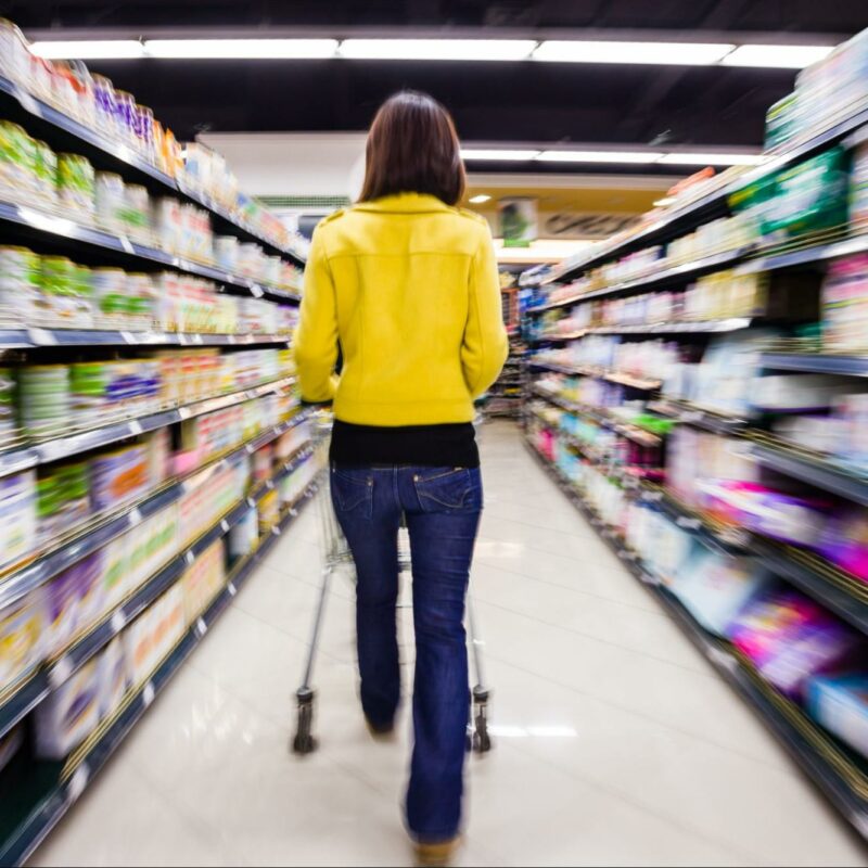 A woman in a yellow jacket and blue jeans shopping, pushing a cart down a grocery store aisle with motion blur.